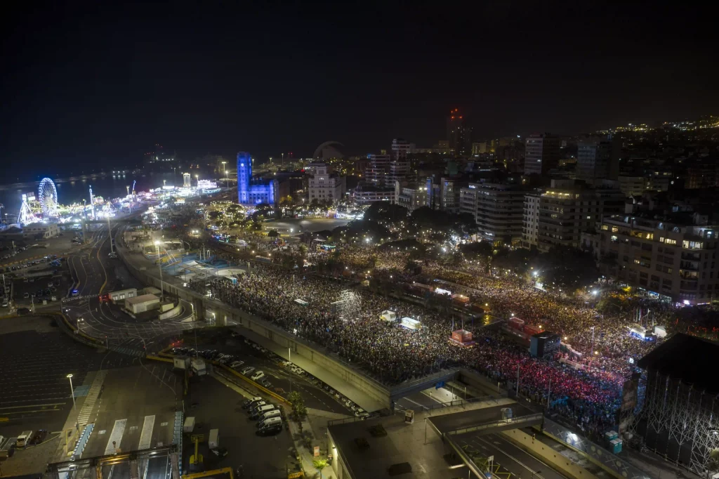 Fiesta nocturna del Carnaval de Tenerife en la Plaza de España, con luces en el escenario, disfraces coloridos y miles de personas.
